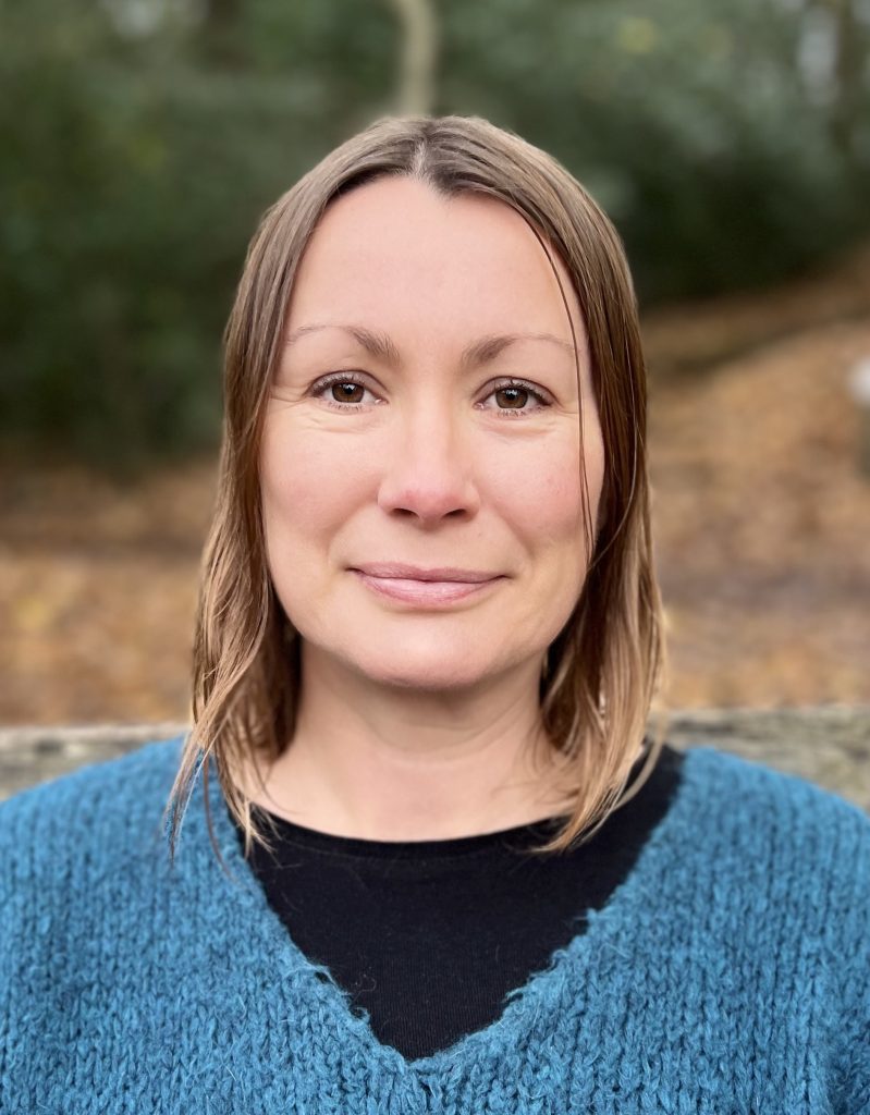A zoomed in head and shoulder photograph of Suzie Bright, with nature backdrop. She is smiling, and has straight shoulder length brown hair. She is wearing a turquoise v-neck jumper.