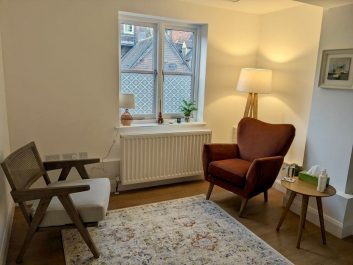Photo of the therapy room in The Old Coach House, Lewes. Two comfortable chairs in between a square window. There is a large faded rug on the floor. There is a floor lamp in the corner casting a soft light across the room. There is a radiator underneath the window.