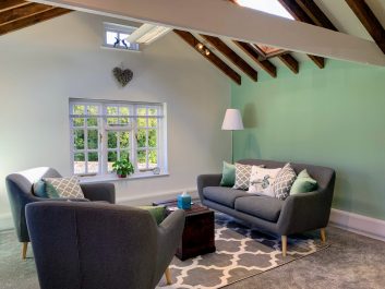 A photo showing the interior of The Buttery therapy room in Glynde, East Sussex. There are 2 grey sofas with lots of cushions facing each other with a coffee table in the middle. Above in the roof are bare oak beams. One of the walls is painted a light green colour. There is a plush grey carpet, with a rug under the coffee table.